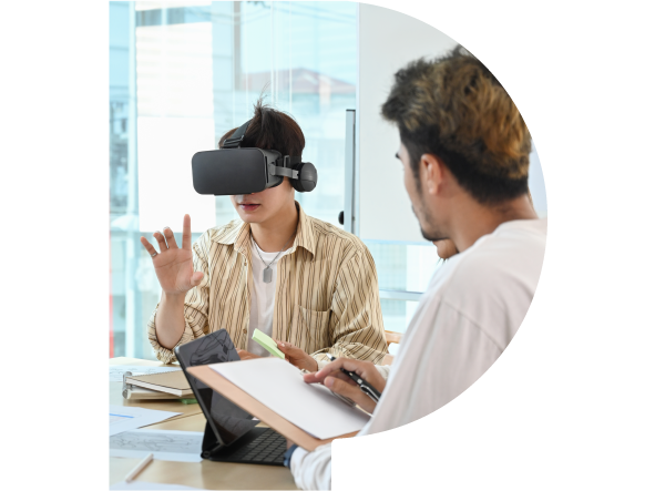 Eine Person trägt eine VR-Brille und zeigt in die Luft. Daneben sitz ein Mann mit Klemmbrett und macht Notizen.