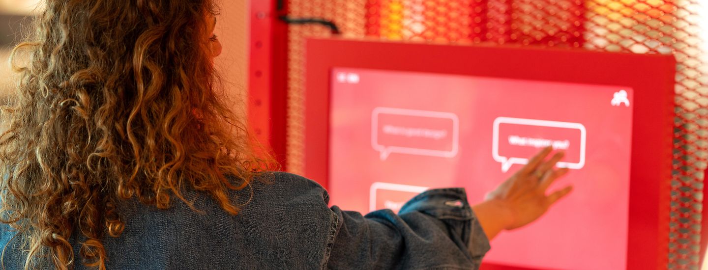Eine Frau steht vor einem Display in einer Ausstellung und berüht mit ihren Fingern den Touchscreen.